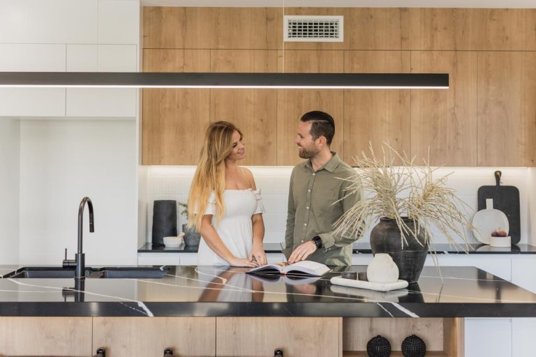 A man and a woman standing in a kitchen looking through a booklet on the counter.